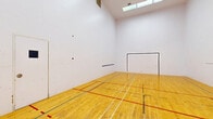 This single-room racquetball court offers a well-appointed, regulation-style playing environment designed for serious recreational use. The court is defined by its polished hardwood floor, finished in a warm natural tone and marked with multiple sets of court lines in red, green, and blue — accommodating a variety of game variations and skill levels.

The surrounding walls are smooth and painted white, providing the consistent, high-contrast surface necessary for gameplay. A clearly marked front wall features a large service box outline, while a horizontal "short line" is visible on the side wall, delineating the key zones of play. The rear wall includes a standard entry door with a small viewing window, allowing observation from outside the court.

The ceiling soars well above the floor, contributing to the open, expansive feel of the space and ensuring unrestricted movement during play. Overhead, skylight panels and recessed ventilation provide natural light supplemented by interior lighting, keeping the court bright and well-ventilated. The overall layout is clean, functional, and purpose-built, making this an excellent facility for both casual players and competitive athletes looking for a dedicated racquetball space.