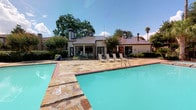 This single-story property is highlighted by a spacious outdoor swimming pool area that serves as a central amenity. The pool is surrounded by a generously sized natural stone deck finished in warm earth tones, providing a durable and visually appealing surface for relaxation and recreation. Several lounge chairs are positioned poolside, offering comfortable outdoor seating beneath the open sky.

The adjacent building features a distinctive standing-seam metal roof in a deep brown tone, complemented by light-colored exterior walls and large glass doors or windows that allow for a visual connection between the interior and the pool area. Architectural details such as a cupola and chimney add character to the roofline. Mature trees, ornamental shrubs, and flowering plants frame the structure, contributing to a lush, well-maintained landscape setting.

The overall ambiance of the space is inviting and resort-like, with the combination of the sparkling turquoise pool, natural stone hardscaping, and verdant surroundings creating an outdoor environment well-suited for leisure. The setting benefits from abundant natural light and open sky views, enhancing the outdoor experience.