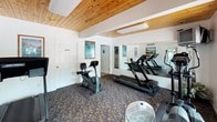 This single-floor fitness center offers a welcoming and functional space designed for cardiovascular exercise and general wellness. The room is defined by its warm, natural wood-plank ceiling, which lends a lodge-like character to the otherwise clean, white-walled interior. Patterned carpeting in earthy tones covers the floor throughout, adding a casual, comfortable feel underfoot.

The space is outfitted with a selection of cardio equipment, including multiple treadmills, an upright stationary bike, an elliptical trainer, and a stair stepper. A large wall-mounted mirror spans one side of the room, visually expanding the space and allowing users to monitor their form during workouts. A mounted television is also present, providing entertainment during exercise sessions.

Natural light filters in through large windows and a glass-paneled door that opens directly to the outdoors, creating a bright and airy atmosphere. Coastal-themed artwork — depicting serene beach and ocean scenes — is displayed on the walls, reinforcing a relaxed, resort-like ambiance.

Additional amenities include a portable fan for supplemental airflow, a wall clock, and an adjacent room accessible through an interior door, suggesting restroom or changing facilities nearby. The overall environment is clean, organized, and well-maintained, making it a practical and inviting space for daily fitness routines.