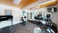 This single-floor fitness center offers a welcoming and functional space designed for cardiovascular exercise and general wellness. The room is defined by its warm, natural wood-plank ceiling, which lends a lodge-like character to the otherwise clean, white-walled interior. Patterned carpeting in earthy tones covers the floor throughout, adding a casual, comfortable feel underfoot.

The space is outfitted with a selection of cardio equipment, including multiple treadmills, an upright stationary bike, an elliptical trainer, and a stair stepper. A large wall-mounted mirror spans one side of the room, visually expanding the space and allowing users to monitor their form during workouts. A mounted television is also present, providing entertainment during exercise sessions.

Natural light filters in through large windows and a glass-paneled door that opens directly to the outdoors, creating a bright and airy atmosphere. Coastal-themed artwork — depicting serene beach and ocean scenes — is displayed on the walls, reinforcing a relaxed, resort-like ambiance.

Additional amenities include a portable fan for supplemental airflow, a wall clock, and an adjacent room accessible through an interior door, suggesting restroom or changing facilities nearby. The overall environment is clean, organized, and well-maintained, making it a practical and inviting space for daily fitness routines.