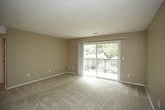 This single-floor residence is designed with a practical, cohesive aesthetic throughout, utilizing warm neutral tones and durable finishes across all rooms. The kitchen is efficiently laid out in a galley-style configuration, featuring white cabinetry, light countertops, wood-look flooring, and white appliances including a stove, dishwasher, and microwave — maximizing utility within a compact footprint.

The living area is generously proportioned, finished with plush wall-to-wall carpeting and neutral beige walls. A standout feature is the large sliding glass door that opens to a private balcony, framing lush green tree views and bringing abundant natural light into the space. A secondary carpeted room, likely a bedroom, mirrors this calm aesthetic and includes a ceiling fan with integrated lighting for added comfort.

The bathroom is well-appointed with wood-look tile flooring, a dark wood vanity with a light countertop, and a full tub-shower combination with white tile surround and chrome fixtures. A grab bar is also present, adding a practical accessibility feature. Throughout the unit, white trim, consistent flooring materials, and a muted color palette create a unified, move-in-ready atmosphere.