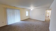 This single-floor space centers around a spacious living area finished with wall-to-wall carpeting in a neutral gray-brown tone, providing a comfortable and cohesive foundation throughout the room. The walls are painted in a soft, light cream color, contributing to a clean and bright atmosphere. A ceiling fan with an integrated light fixture is centrally positioned, offering both overhead illumination and air circulation year-round. One wall features a large sliding glass door dressed with full-length vertical blinds, allowing for adjustable natural light and potential outdoor access. An additional window on the adjacent wall further enhances the natural light entering the space. Clean white baseboards run along the perimeter of the room, adding a crisp, finished detail to the interior. Multiple electrical outlets are positioned along the walls, offering practical functionality throughout the room. The overall layout is open and unobstructed, providing flexibility for a variety of furniture arrangements and uses. The space presents a straightforward, functional design with a neutral palette well-suited for personalization.