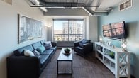 This 4-bedroom, 2-bathroom unit showcases an industrial-modern aesthetic throughout its single-floor layout. Exposed ductwork and concrete ceilings define the architectural character, complemented by rich hardwood flooring in the main living areas and durable carpet in the bedrooms.

The open-concept kitchen is well-appointed with light wood cabinetry, dark granite countertops, black appliances including a range and built-in microwave, and a central island with bar seating — ideal for both cooking and casual dining. The adjoining living area is bright and spacious, featuring large windows that frame urban views of the surrounding cityscape.

Each of the four bedrooms offers a private, functional layout with carpeted floors and room for a bed and dedicated study desk, making them well-suited for student living. Several bedrooms benefit from large windows with open city views. The two full bathrooms are clean and simply designed, each featuring a shower and essential fixtures.

The unit's industrial design elements — including visible pipes and ductwork — lend a loft-inspired feel, while the teal accent walls and generous natural light throughout add warmth and visual interest to the overall space.