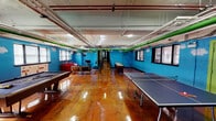 Located at 5445 S Ingleside Ave in Chicago's Hyde Park neighborhood, this single-floor recreational space is designed for active social engagement and entertainment. The interior is immediately striking, defined by bold sky-blue painted brick walls adorned with whimsical hand-painted cloud murals and colorful video game character decals, creating a lively and playful atmosphere throughout.

The highly polished epoxy-coated concrete flooring in rich amber and mahogany tones reflects light beautifully across the open layout, lending an industrial yet vibrant character to the space. Exposed overhead piping in contrasting green and white runs along the ceiling, complementing the industrial aesthetic while adding a pop of color.

The game area is generously sized, accommodating a full pool table and a regulation ping-pong table with ample room to play comfortably. A dedicated lounge corner features a bold red sofa and additional seating arranged around a flat-screen television, offering a relaxed retreat within the activity-focused environment.

Multiple windows line the walls, allowing natural light to filter in. The space is served by fluorescent overhead lighting, ensuring bright, consistent illumination throughout. A green-painted entry door adds another vibrant accent. Overall, this energetic space is well-suited for community gatherings, recreation, and social events.