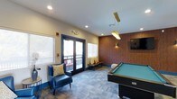 This inviting game room is thoughtfully designed to serve as a social hub for recreation and relaxation. At the heart of the space sits a full-sized pool table beneath a striking gold pendant light, creating a natural focal point that draws the eye. A warm wood-paneled accent wall adds depth and character, complemented by a wall-mounted flat-screen TV, making the room equally suited for both active play and casual viewing. Recessed lighting throughout ensures the space is well-lit, while a bold blue armchair with a patterned throw pillow and a bench with coordinating cushions offer comfortable seating options along the perimeter. The patterned carpet tiles add texture underfoot and contribute to the room's contemporary aesthetic. Large windows and a set of navy-trimmed glass French doors flood the interior with natural light and open directly onto an outdoor balcony, extending the living area and providing a seamless indoor-outdoor connection. The overall ambiance strikes a balance between energetic and laid-back, making this game room a versatile amenity space well-suited for socializing, entertaining, and unwinding.
