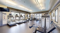 This well-equipped fitness center at 1311 Holling Pl in Frederick, Maryland, offers a comprehensive workout environment within a single, open-plan room. The space features durable rubber flooring throughout, warm beige walls, and a tray ceiling with light blue accents, lending the room a clean, modern aesthetic. Overhead fluorescent lighting is supplemented by abundant natural light streaming through multiple large windows dressed with slatted blinds, creating a bright and energizing atmosphere.

The cardio section is prominently anchored by a row of treadmills and an elliptical trainer, all equipped with individual display screens. Strength training is well represented with multiple cable and weight machines, including a multi-function cable station, leg press, and seated strength machines. A dedicated free weights area features a full rack of dumbbells alongside a weight bench, catering to a range of training styles. Exercise accessories such as stability balls are also available.

A large wall mirror along one side of the room enhances the sense of space and supports proper form during workouts. Exposed ductwork running along the upper walls contributes an industrial design element. Posted fitness center rules and a sanitization station reflect a commitment to cleanliness and order. The space is accessible via a key-entry door, ensuring a secure environment for users.