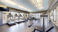 This well-equipped fitness center at 1311 Holling Pl in Frederick, Maryland, offers a comprehensive workout environment within a single, open-plan room. The space features durable rubber flooring throughout, warm beige walls, and a tray ceiling with light blue accents, lending the room a clean, modern aesthetic. Overhead fluorescent lighting is supplemented by abundant natural light streaming through multiple large windows dressed with slatted blinds, creating a bright and energizing atmosphere.

The cardio section is prominently anchored by a row of treadmills and an elliptical trainer, all equipped with individual display screens. Strength training is well represented with multiple cable and weight machines, including a multi-function cable station, leg press, and seated strength machines. A dedicated free weights area features a full rack of dumbbells alongside a weight bench, catering to a range of training styles. Exercise accessories such as stability balls are also available.

A large wall mirror along one side of the room enhances the sense of space and supports proper form during workouts. Exposed ductwork running along the upper walls contributes an industrial design element. Posted fitness center rules and a sanitization station reflect a commitment to cleanliness and order. The space is accessible via a key-entry door, ensuring a secure environment for users.