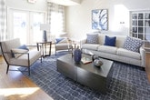 This thoughtfully designed single-floor space makes an immediate impression with its dramatic great room, where soaring ceilings rise to a clerestory window bank that floods the interior with abundant natural light. Warm hardwood floors run throughout, tying each area together with a sense of cohesion and continuity.

The main living area is generously proportioned, anchored by a comfortable neutral linen sofa accented with blue geometric and solid throw pillows. A textured navy area rug grounds the seating arrangement, while recessed lighting and large framed abstract artwork in blues and whites contribute to the room's refined yet approachable atmosphere. A stone fireplace is visible in the background, adding architectural character and a cozy focal point.

Adjacent to the living area, a dedicated coffee bar brings both function and personality to the space. A bold chalkboard-painted accent wall features floating wood shelves stocked with coffee essentials, playful handwritten phrases, and decorative items. White cabinetry with granite countertops below provides ample storage and prep space.

A bright corridor or transition area connects the rooms, styled with a sleek geometric metal console table, curated ceramic décor in complementary blue tones, and large-scale abstract artwork. Plaid drapery panels and plantation shutters on the windows offer light control while maintaining a polished aesthetic. Together, these elements create a space that is both visually striking and highly functional.