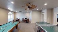 This well-appointed billiards room is designed for leisure and entertainment, offering a variety of game options within a single, open space. At the heart of the room sits a classic pool table with a rich wood finish and green felt surface, ready for competitive play. Across the room, a full-sized ping pong table provides an additional layer of recreational fun. A dedicated card and game table, surrounded by wicker-style cushioned chairs, offers a comfortable spot for board games or casual conversation. The room features neutral-toned carpeting that adds warmth underfoot, while the light gray walls create a clean, inviting backdrop. Recessed lighting throughout ensures ample illumination for all activities, complemented by a distinctive tropical-style ceiling fan that adds a relaxed, resort-like ambiance to the space. A cue rack is conveniently mounted near the card table, keeping accessories organized and within reach. The room is well-ventilated with ceiling vents and benefits from natural light through a window along the side wall, contributing to a bright and welcoming atmosphere. Overall, this billiards room is a thoughtfully equipped recreational space suited for guests and residents of all ages.