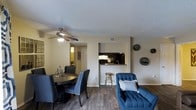 This single-floor residence offers a well-organized, open-concept layout that seamlessly connects the living, dining, and kitchen areas into one cohesive living space. The flooring throughout is a rich hardwood-style material that adds warmth and a contemporary feel underfoot. The dining area comfortably accommodates a round table with upholstered chairs, accented by a ceiling fan with integrated lighting that provides both comfort and illumination. Decorative wall accents add a curated touch to the neutral-toned walls. The kitchen is positioned at the rear of the open space and features dark cabinetry, under-cabinet lighting, and a raised breakfast bar with barstool seating — ideal for casual dining or entertaining. A pass-through design between the kitchen and living area maintains the open flow of the space. The living area is furnished with a plush, tufted blue accent chair, contributing a bold pop of color against the neutral palette. A textured popcorn ceiling is consistent throughout. The overall ambiance is modern and functional, with a layout that maximizes the use of space while maintaining a comfortable and inviting atmosphere.