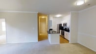 This single-floor residence features an open-concept design that integrates the living area and kitchen into one cohesive space. The living area is finished with plush carpet flooring, while the kitchen transitions to hardwood-style flooring, creating a natural visual distinction between the two zones. Crown molding runs along the ceiling throughout, adding a refined architectural detail to the otherwise clean, neutral interior.

The kitchen is well-appointed with white cabinetry, dark granite countertops, and stainless steel appliances, including a refrigerator, microwave, and range. A peninsula with a built-in sink extends into the space, offering additional prep area and serving as a functional divider between the kitchen and living room. The layout allows for easy interaction between both areas.

A hallway visible from the main living space leads toward the entry door, finished with tile flooring and flanked by closet space, providing a defined transition zone upon entry. Flush-mount ceiling lighting illuminates both the kitchen and hallway. The overall palette of soft whites, warm grays, and neutral tones gives the space a clean, move-in-ready ambiance suited for everyday living.