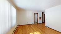 This single-floor residence offers a well-organized layout with a variety of distinct living spaces, each with its own character and function. The main living area is immediately inviting, featuring gleaming hardwood floors that extend throughout much of the unit and large windows dressed with vertical blinds that allow generous natural light to fill the room. The bright, neutral walls complement the warm wood tones, creating an open and airy atmosphere.

The kitchen is thoughtfully equipped with warm oak cabinetry both above and below the countertops, providing ample storage. A double stainless steel sink sits beneath a small interior window, and the space includes a dishwasher and full-size refrigerator. Light tile flooring keeps the kitchen practical and easy to maintain.

The bedroom is a quiet, carpeted retreat featuring a ceiling fan, a wood-trimmed window with blinds, and baseboard heating — offering comfort through all seasons.

The full bathroom includes a tiled tub-and-shower combination, a pedestal-style sink, a wood vanity, and tile flooring, accessed through a classic wood-framed doorway. Additional features throughout the unit include floor vents for heating and cooling, consistent warm-toned wood trim on all doors and windows, and a cohesive neutral color palette that gives the space a clean, move-in-ready feel.