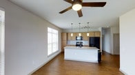 This single-story space in Texas showcases an open-concept design that seamlessly connects the living area and kitchen. The flooring throughout is warm-toned wood-look hardwood, lending a cohesive and inviting feel to the interior. A ceiling fan with a wood-finish complements the neutral white walls, promoting airflow and comfort.

The kitchen is a standout feature, equipped with medium-toned wood cabinetry that provides ample storage. Black appliances, including a refrigerator with an ice dispenser, a range, and a built-in microwave, are neatly integrated. A central island with a light granite-style countertop and a sink serves as a functional workspace and gathering point. Above the island, a trio of pendant lights add a stylish focal point, while additional track lighting illuminates the surrounding area.

The adjacent living space benefits from a large window dressed with horizontal blinds, allowing natural light to fill the room. The open layout creates a sense of spaciousness, making the area well-suited for both everyday living and entertaining. The overall ambiance is clean, modern, and practical.