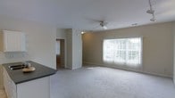 This single-floor residence offers a clean, functional layout designed for comfortable everyday living. The open-concept living area is a standout feature, characterized by soft, plush carpeting underfoot and neutral-toned walls that create a calm, welcoming atmosphere. A ceiling fan is centrally positioned overhead, providing both practical ventilation and a finishing touch to the room. A large window dressed with vertical blinds allows generous natural light to flow in, brightening the space throughout the day and offering views of the surrounding outdoor greenery. The living area connects seamlessly to the kitchen, which is partially visible and features dark countertops contrasted by white cabinetry, lending a classic and timeless aesthetic. A hallway visible from the main living space suggests additional rooms beyond, hinting at a thoughtful floor plan that balances open communal areas with more private spaces. Clean white baseboards frame the carpeted floors throughout, adding a crisp, polished detail to the overall interior. The space presents a straightforward, move-in-ready environment with a light, airy ambiance well-suited for a range of lifestyles.