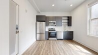 This contemporary single-floor unit offers a thoughtfully designed layout with modern finishes throughout. The open-plan kitchen is a standout feature, showcasing charcoal gray cabinetry that extends from floor to ceiling, providing ample storage. A full suite of stainless steel appliances — including a French door refrigerator, electric range, over-the-range microwave, and dishwasher — is complemented by a clean white tile backsplash and light-toned countertops. Light wood-look flooring runs throughout the space, adding warmth and continuity. Recessed lighting and white walls create a bright, airy atmosphere in the kitchen and living area. The bedroom benefits from generous natural light through a large multi-pane window, and features double doors leading to a sizable closet. The bathroom is finished in a crisp, all-white palette with large-format floor tiles and a frameless glass-enclosed walk-in shower equipped with both a fixed showerhead and a handheld fixture. A grab bar and clean, minimalist fixtures add functional detail. Overall, the unit presents a cohesive modern aesthetic, combining practical storage solutions with clean design elements throughout.
