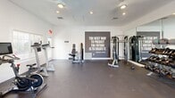 This compact yet fully outfitted fitness center offers a comprehensive workout environment within a single, efficiently designed exercise room. The space features dark rubber flooring throughout, providing a durable and functional surface suited for a wide range of training activities. Recessed lighting and ceiling fans maintain a bright, comfortable atmosphere, while large wall-mounted mirrors along one side visually expand the room and assist with form and technique during workouts.

The cardio zone is equipped with treadmills, elliptical machines, and a stationary cycling bike, all positioned near a window with natural light filtering through slatted blinds. A wall-mounted flat-screen television accompanies the cardio area for entertainment during workouts. On the strength side, a multi-station cable machine anchors the equipment selection, complemented by a well-stocked dumbbell rack ranging across multiple weight increments, kettlebells, and medicine balls. A BOSU balance trainer adds versatility for functional fitness training.

Motivational quotes — including an Abraham Lincoln attribution reading "The best way to predict the future is to create it" — are displayed prominently on a bold dark accent wall, lending energy and inspiration to the space. French doors open toward what appears to be an outdoor pool area, offering a refreshing view and natural ventilation. The room is accessible via a dedicated entrance with clearly marked exit signage.
