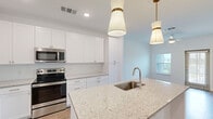 Located at 7785 Griffon Ln in Vero Beach, Florida, this single-story, one-bedroom, two-bathroom home offers a thoughtfully designed layout that maximizes its compact footprint. The open-concept kitchen serves as the centerpiece of the living space, showcasing white shaker cabinetry with gold hardware, light granite countertops, and a large island with an undermount sink. Stainless steel appliances — including a French door refrigerator, electric range, and built-in microwave — complement the clean, contemporary aesthetic. Two pendant lights with gold accents hang above the island, adding a refined touch. Wood-look flooring runs through the main living and kitchen areas, providing warmth and continuity. The living area is bright and open, flowing naturally from the kitchen. The primary bedroom features plush carpeting, a ceiling fan, dual windows that invite natural light, and direct access to a walk-in closet and a private bathroom with granite vanity. A second full bathroom serves the remainder of the home. White walls and recessed lighting throughout create a clean, airy ambiance well-suited to Florida living.