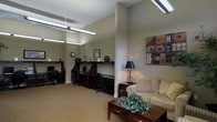 This single-floor space is thoughtfully designed to serve dual purposes, seamlessly blending a relaxed lounge environment with a practical business center area. The lounge section features a cushioned sofa, a wooden coffee table adorned with greenery, and a side table with a classic table lamp, creating a warm and inviting atmosphere. Neutral-toned walls and wall-to-wall carpeting in earthy tones contribute to a calm, comfortable ambiance throughout.

Adjacent to the lounge, the workstation area is equipped with multiple individual computer desks separated by partitioned dark wood dividers, each station featuring its own monitor and task chair. Overhead fluorescent strip lighting ensures ample illumination across the workspace, making it well-suited for productivity. A wall clock and decorative accents, including a motivational wall sign, add personality to the area.

The space is unified by its consistent warm color palette and complementary furnishings. Decorative artwork featuring bold geometric patterns provides a visual focal point on the lounge side, tying together the aesthetic of the room. Overall, the layout is both functional and welcoming, offering residents or visitors spaces for both leisure and focused work.