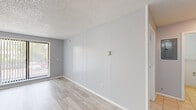 This one-bedroom, one-bathroom unit in Rensselaer, NY offers a well-organized single-floor layout that efficiently incorporates a living room, kitchen, bedroom, and bathroom. The living room serves as the central gathering space, featuring light wood-look flooring, neutral gray panel walls, and a textured ceiling. A large sliding window with vertical blinds allows natural light to fill the room while providing views of the surrounding greenery, and a baseboard heating system runs along the perimeter wall. The bedroom is a comfortable, dedicated space that mirrors the living area's clean aesthetic with the same flooring and neutral wall tones, complemented by additional baseboard heating. The kitchen is equipped with light blue cabinetry offering ample upper and lower storage, dark granite-style countertops, a stainless steel sink, a stainless steel refrigerator, and a stainless steel range with an overhead ventilation hood. Light tile flooring grounds the kitchen area. The bathroom includes a full bathtub with a surround enclosure. Throughout, the consistent use of light tones and wood-look flooring creates a cohesive, clean ambiance suited for practical everyday living.