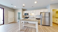 This single-floor residence showcases a clean, contemporary design with a neutral color palette and light-toned finishes throughout. The open-concept kitchen serves as the focal point, outfitted with white shaker-style cabinetry, granite countertops, and a full suite of stainless steel appliances including a range, over-the-range microwave, dishwasher, and refrigerator. A freestanding island with a matching granite surface provides additional workspace and casual dining. Light wood-look luxury vinyl plank flooring extends across the kitchen and living areas, lending warmth and continuity to the space. Recessed lighting illuminates the main living area, which opens toward a sliding glass door that connects to a balcony, allowing natural light to flow in. The bedrooms are finished with plush neutral-toned carpet and feature individual windows that bring in additional daylight. The bathroom is well-appointed with a granite-topped vanity featuring white cabinetry, a soaking tub with shower surround, and matching wood-look flooring. The overall ambiance is bright, clean, and move-in ready, with modern finishes creating a cohesive and functional living environment.