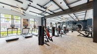 Located at 3175 Corporate Center Pkwy in Kissimmee, Florida, this well-appointed fitness center offers a generously sized, single-floor workout facility designed with both functionality and aesthetics in mind. The space features a striking industrial-modern aesthetic, highlighted by a bold coffered ceiling with dark steel beam accents, integrated linear lighting, and exposed ductwork that adds an urban, loft-like character.

Large black-framed floor-to-ceiling windows line the perimeter, flooding the interior with abundant natural light and offering views of the lush, palm-tree-lined outdoor surroundings. The main workout floor is fully equipped with a row of treadmills and cardio machines positioned along the windowed wall, multiple Hoist strength machines, adjustable benches, a full dumbbell rack, and cable equipment—catering to a wide range of fitness routines.

A dedicated free weights area features arched, backlit mirror panels set against a deep charcoal accent wall, creating a sophisticated focal point. Additional visual interest comes from a textured hexagonal wall panel in dark tones, decorative blueprint-inspired wall art, and motivational signage. A storage closet and equipment station with cubbies provide practical utility. The neutral-toned carpet flooring throughout ties together the space's refined, high-end gym aesthetic.