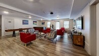 Located at 38 Greenleaves Dr in Hadley, MA, this single-floor communal space offers a well-appointed lounge environment designed for social interaction and recreation. The open layout is unified by durable wood-look flooring that runs throughout, lending warmth and visual continuity to the entire area.

The main lounge zone is furnished with a mix of bold red accent chairs and patterned geometric armchairs arranged around small coffee tables, creating multiple comfortable conversation areas. A wall-mounted flat-screen TV positioned above a rustic wood media console serves as a central focal point for the seating arrangement. Colorful abstract artwork displayed along the walls adds personality and energy to the space.

Toward the back of the room, a game table provides a dedicated recreational area, complemented by a bar-height counter with red stools—ideal for casual seating or activity. A stainless steel refrigerator is also present, adding functional convenience. Natural light filters in through multiple windows along the perimeter, while flush-mounted ceiling fixtures ensure consistent interior illumination. An exterior door with sidelight panels offers direct access to the outdoors. Together, these elements create a lively, multi-functional communal environment suited for group activities and relaxed gatherings.