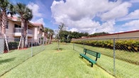 This outdoor dog park offers a dedicated, fully enclosed space designed for pets and their owners to enjoy fresh air and open greenery. The park features a spacious, well-kept lawn enclosed by a chain-link fence, providing a secure environment for dogs to roam freely. A green metal bench is conveniently positioned within the enclosure, offering a comfortable resting spot for owners while their pets play. The grounds are lush and neatly maintained, with vibrant green grass throughout. The surrounding landscape is rich with mature trees, tropical palm trees, and dense hedging, lending a natural and serene atmosphere to the space. The park benefits from abundant natural light and open sky views, with picturesque clouds adding to the inviting outdoor ambiance. A multi-story residential building with a terracotta tile roof is visible in the background, situating the park within a well-established community setting. The combination of secure fencing, ample open space, seating, and verdant surroundings makes this dog park a functional and pleasant amenity for residents and their pets.