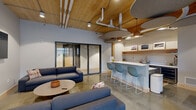 Located at 300 N Cherapa Place in Sioux Falls, South Dakota, this thoughtfully designed communal lounge space offers a dynamic and inviting atmosphere ideal for socializing and relaxation. The open-plan layout is defined by warm natural wood plank ceilings accented by exposed ductwork and structural timber posts, creating an industrial-modern aesthetic that feels both polished and approachable. Polished concrete floors run throughout, lending a clean, contemporary foundation to the space.

The lounge area features comfortable blue sectional and gray sofas arranged around a round coffee table, complemented by a wall-mounted flat-screen television — perfect for casual gatherings. A built-in kitchenette with a white countertop bar and teal bar stools provides a convenient refreshment station, complete with open shelving for display. Suspended acoustic panels in soft gray tones hang from the ceiling, aiding sound quality while adding a sculptural design element.

Large floor-to-ceiling steel-framed windows and glass sliding doors flood the space with natural light and offer direct views into a landscaped outdoor courtyard. A dedicated dining nook with small round tables and modern chairs sits adjacent to these windows. A foosball table adds a recreational touch, rounding out this vibrant, multi-functional community space.