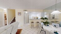 This two-story residence is designed with a bright, open layout that maximizes the sense of space throughout. The main floor centers around a cohesive living, dining, and kitchen area finished with large white ceramic tiles that reflect natural light from multiple windows. The living room features white leather seating, decorative area rugs, and ornate window treatments that add warmth to the otherwise crisp palette. A full-wall mirror in the dining area visually expands the space, which accommodates a round dining table and chairs alongside a pendant light fixture.

The kitchen is defined by its peninsula with granite countertops, white cabinetry extending to the ceiling, and white appliances including a refrigerator and range. Two bar stools at the peninsula create a casual seating option. Louvered bifold doors conceal a laundry area conveniently located adjacent to the kitchen. A full bathroom on the main floor includes a shower-tub combination and tile flooring.

Upstairs, the primary bedroom offers a textured ceiling, ceiling fan, natural light from a window with garden views, and access to a private hallway leading to a full bathroom with a soaking tub. The upper level maintains the same neutral, warm color palette found throughout the home.