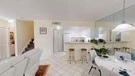 This two-story residence is designed with a bright, open layout that maximizes the sense of space throughout. The main floor centers around a cohesive living, dining, and kitchen area finished with large white ceramic tiles that reflect natural light from multiple windows. The living room features white leather seating, decorative area rugs, and ornate window treatments that add warmth to the otherwise crisp palette. A full-wall mirror in the dining area visually expands the space, which accommodates a round dining table and chairs alongside a pendant light fixture.

The kitchen is defined by its peninsula with granite countertops, white cabinetry extending to the ceiling, and white appliances including a refrigerator and range. Two bar stools at the peninsula create a casual seating option. Louvered bifold doors conceal a laundry area conveniently located adjacent to the kitchen. A full bathroom on the main floor includes a shower-tub combination and tile flooring.

Upstairs, the primary bedroom offers a textured ceiling, ceiling fan, natural light from a window with garden views, and access to a private hallway leading to a full bathroom with a soaking tub. The upper level maintains the same neutral, warm color palette found throughout the home.