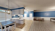 Located in Madison, Alabama, this single-floor space offers a well-organized, open-concept layout encompassing a kitchen, dining room, and living area. A consistent and striking blue-and-white color palette ties the entire space together, with rich blue walls accented by white wainscoting, crown molding, and trim throughout.

The kitchen is functional and bright, featuring white cabinetry, white countertops, a stainless steel sink, track lighting, and tile flooring. Multiple doorways provide convenient access to adjoining areas. Adjacent to the kitchen, the dining room is generously lit by large windows with transom panels and white blinds that invite natural light and outdoor views. A modern globe pendant light hangs above the dining table, adding a contemporary touch to the classic trim details.

The living area transitions seamlessly from the tiled kitchen zone to wood-look laminate flooring and includes a spacious lounge and a recreation zone complete with a pool table beneath a hanging pendant light and a statement oversized wall clock. A separate seating area features a sectional sofa, ceiling fan, and abstract wall art. The textured ceilings and cohesive crown molding throughout lend a polished, finished feel. Overall, this space combines practicality with a lively, welcoming atmosphere well-suited for community or social use.
