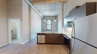 This thoughtfully designed loft-style unit masterfully bridges industrial character with modern living. The open-concept kitchen serves as a centerpiece, outfitted with dark wood cabinetry, stone countertops, a full suite of stainless steel appliances — including a refrigerator, range, built-in microwave, and dishwasher — and stylish pendant lighting. A peninsula with a undermount sink provides both functional workspace and a natural division between the kitchen and the living area. Original exposed brick walls are a defining architectural feature throughout the space, lending warmth, texture, and historic character. Soaring ceilings with exposed ductwork reinforce the industrial loft aesthetic, while light hardwood-style flooring runs through the main living areas, adding a clean, contemporary contrast. The bedroom showcases a striking full brick wall with a tall arched window that frames urban views, bringing in abundant natural light. Soft carpet underfoot provides comfort in this private retreat. The bathroom is finished with a dark wood vanity, stone countertop, large mirror, and light hardwood-style flooring, maintaining the cohesive design language found throughout the unit. This space offers a compelling blend of historic charm and refined modern upgrades.