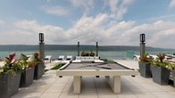 Located atop 1077 Warburton Avenue in Yonkers, NY, this rooftop deck offers an exceptional amenity space designed for relaxation and recreation with sweeping, unobstructed views of the Hudson River and the lush Palisades cliffs beyond. The outdoor deck is outfitted with a full-sized outdoor billiards table as a central focal point, surrounded by stylish lounge furniture and sun chairs, all set against a transparent glass railing that preserves the breathtaking panoramic scenery. Large planter beds add a touch of greenery, softening the clean, modern hardscape of the tiled deck surface. Tall, contemporary post lighting is positioned throughout the outdoor space, ensuring ambiance during evening gatherings. Access to the rooftop is provided through a bright, well-appointed interior lobby featuring light herringbone-patterned flooring, soft neutral walls, and modern wall-mounted sconces. A stainless steel elevator serves the rooftop level directly, and oversized double glass doors with transom windows frame a preview of the outdoor deck and river views beyond. A vibrant large-scale coastal artwork adorns the lobby wall, reinforcing the waterfront character of the space. Together, the interior vestibule and expansive outdoor deck create a polished, resort-inspired rooftop experience.