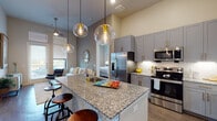 This well-appointed single-floor residence showcases a thoughtfully designed open-concept layout with soaring ceilings that create an airy, expansive feel throughout. The kitchen serves as the heart of the home, featuring gray shaker-style cabinetry with gold hardware, granite countertops, a large center island with bar seating, and a full suite of stainless steel appliances including a refrigerator with ice maker, range, and built-in microwave. Decorative globe pendant lights hang above the island, adding a warm, modern touch. Hardwood-style flooring flows seamlessly through the main living areas, connecting the kitchen to a comfortable living room highlighted by tall windows and a glass-paned door that invite abundant natural light and outdoor views. The bedrooms offer a calm retreat, each with large windows, neutral tones, and hardwood-style floors. The primary suite includes an en suite bathroom with a granite-topped double vanity, gray cabinetry, and direct access to a spacious walk-in closet outfitted with built-in shelving and hanging rods. Throughout the residence, consistent finishes — including granite surfaces, gray cabinetry, and gold accents — lend a cohesive, contemporary aesthetic.
