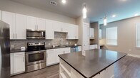 This single-story space is designed with a contemporary aesthetic, offering a cohesive blend of modern finishes and functional layouts. The kitchen serves as a central highlight, featuring crisp white shaker-style cabinetry extending to the ceiling, dark countertops, and a textured stone tile backsplash that adds visual depth. A full suite of stainless steel appliances — including a French door refrigerator, electric range, over-the-range microwave, and dishwasher — lines the perimeter walls, while a central island with matching dark countertop provides additional prep and seating space. Cylindrical pendant lights hang above the island, adding a refined touch. Wood-look plank flooring runs through the kitchen and adjoining living areas, providing a warm, durable surface. A dedicated built-in workspace or bar area with open shelving is visible off the kitchen. The living area features recessed lighting, a ceiling fan, and a wide window fitted with blinds that allows natural light to fill the room. Secondary bedroom spaces are carpeted with neutral tones and include windows with blinds. A walk-in closet with built-in shelving and a hanging rod offers organized storage. Overall, the space maintains a clean, neutral palette throughout.