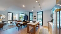This contemporary single-floor residence showcases a thoughtfully designed open-concept layout that seamlessly connects the living, dining, and kitchen areas. The living room is bathed in natural light through expansive floor-to-ceiling windows with sleek black frames, offering urban views of the surrounding cityscape. Light hardwood flooring runs throughout the main living spaces, complementing the clean, modern aesthetic.

The kitchen features a two-tone design with white upper cabinetry and warm wood lower cabinets, white subway tile backsplash, quartz countertops, and stainless steel Bosch appliances including a range, microwave, and dishwasher. A freestanding island provides additional prep and dining space.

The primary bedroom offers dual windows with sheer draping curtains and access to an en-suite bathroom finished with large-format light tile flooring, a wood-grain vanity with white quartz countertop, an illuminated mirror, and a glass-enclosed walk-in shower with rich dark tile surround. A second bedroom features a walk-in closet and its own access to a full bathroom appointed with a soaking tub and similarly styled dark tile shower surround. The overall ambiance is refined and urban-contemporary throughout.