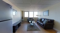 This single-floor space offers a well-appointed living area designed for comfortable, everyday use. The room is anchored by warm hardwood flooring that extends throughout, complemented by neutral-toned walls that create a clean, understated backdrop. A drop tile ceiling adds a practical, finished look to the interior.

The living area is generously proportioned, accommodating a dark leather sofa and a cushioned armchair arranged around a glass-top oval coffee table. A patterned area rug in muted tones grounds the seating arrangement, adding texture and visual warmth. A low-profile media console sits along one wall, providing functional storage and entertainment space.

Natural light enters through a row of horizontally aligned windows that span one wall, offering elevated views of a city skyline — a notable feature that adds an urban character to the space. Framed artwork on the walls and accent pieces, including a floor lamp and decorative side tables, contribute to the room's lived-in, functional aesthetic.

Overall, the space presents a straightforward and practical layout, with a balance of comfort and utility suited to daily residential use.