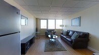 This single-floor space offers a well-appointed living area designed for comfortable, everyday use. The room is anchored by warm hardwood flooring that extends throughout, complemented by neutral-toned walls that create a clean, understated backdrop. A drop tile ceiling adds a practical, finished look to the interior.

The living area is generously proportioned, accommodating a dark leather sofa and a cushioned armchair arranged around a glass-top oval coffee table. A patterned area rug in muted tones grounds the seating arrangement, adding texture and visual warmth. A low-profile media console sits along one wall, providing functional storage and entertainment space.

Natural light enters through a row of horizontally aligned windows that span one wall, offering elevated views of a city skyline — a notable feature that adds an urban character to the space. Framed artwork on the walls and accent pieces, including a floor lamp and decorative side tables, contribute to the room's lived-in, functional aesthetic.

Overall, the space presents a straightforward and practical layout, with a balance of comfort and utility suited to daily residential use.