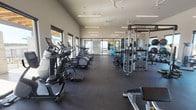 This well-appointed fitness center offers a thoughtfully designed single-floor layout that accommodates a wide range of workout disciplines. The space features soaring vaulted ceilings with sleek linear LED lighting and ceiling fans that maintain an open, airy atmosphere throughout. Large picture windows line multiple walls, flooding the interior with natural light and providing expansive views of the surrounding landscape and outdoor amenity areas.

The cardio zone is equipped with multiple treadmills, elliptical machines, and stationary bikes arranged along the windowed perimeter, allowing users to exercise with scenic outdoor views. The strength training area includes a robust selection of cable machines, selectorized equipment, squat racks, adjustable benches, and a full dumbbell rack, all positioned alongside floor-to-ceiling mirrors that enhance spatial depth. A dedicated functional training zone features stability and medicine balls, providing space for stretching and bodyweight exercises.

Dark rubber flooring throughout offers both durability and comfort underfoot. The entrance area features sleek black-framed glass doors, adding a contemporary architectural touch. The overall design is clean, modern, and highly functional, creating a professional gym environment suitable for a full spectrum of fitness routines.