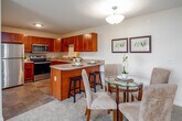 This single-floor residence offers a thoughtfully designed open-concept layout that seamlessly connects the living, dining, and kitchen areas. The living room is bright and inviting, featuring plush carpeting, a wall-mounted TV, and a large sliding glass door that opens to a private balcony with views of the surrounding neighborhood. A ductless mini-split climate control unit ensures year-round comfort. The dining area accommodates a glass-top round table with upholstered chairs, positioned conveniently adjacent to the kitchen peninsula with bar seating. The kitchen is well-equipped with rich cherry wood cabinetry, granite-style countertops, stainless steel appliances including a refrigerator, range, microwave, and dishwasher, and durable tile flooring. Two carpeted bedrooms provide comfortable retreats, each with windows that allow ample natural light. The primary bedroom features dual windows with views to the outdoors, while the secondary bedroom includes a dedicated desk area. The full bathroom is finished with tile flooring, a wood vanity, a framed mirror, a glass-enclosed shower, and accessibility-friendly grab bars. Throughout the residence, neutral tones and consistent finishes create a cohesive and welcoming ambiance.
