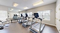 Located at 201 Eaton Ridge Dr in Northfield, Ohio, this dedicated fitness center offers a well-equipped exercise space designed to support a well-rounded workout routine. The room features durable speckled flooring suited for high-traffic athletic use, and is illuminated by recessed flush-mount fluorescent ceiling fixtures that ensure bright, consistent lighting throughout.

The space is outfitted with multiple True-brand treadmills, an elliptical trainer, a stationary recumbent bike, and a multi-function cable weight machine, providing a comprehensive range of cardiovascular and strength-training options. A free-weight rack with dumbbells and a workout bench round out the strength-training offerings. A wall-mounted flat-screen television adds entertainment value for users during cardio sessions.

Large floor-to-ceiling mirrors line one wall, visually expanding the space and allowing users to monitor their form during exercise. Multiple windows fitted with white plantation-style blinds invite natural light while maintaining privacy, contributing to a bright and open atmosphere. The neutral gray-toned walls and white trim create a clean, focused environment. A glazed interior door provides access to the adjoining area. Overall, the fitness center is a compact yet thoughtfully arranged space that maximizes functionality for residents seeking convenient on-site exercise facilities.