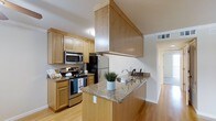 This single-floor 2-bedroom, 2-bathroom residence combines practical design with modern finishes to create a comfortable and inviting living environment. The kitchen serves as a standout feature, showcasing warm honey-toned wood cabinetry that extends to the ceiling, providing generous storage throughout. A peninsula with a granite countertop offers additional prep space and doubles as a casual dining or gathering area. Stainless steel appliances, including a range, microwave, and refrigerator, add a contemporary touch and are well-integrated into the cabinetry layout. A built-in range hood encased in matching wood paneling adds an architectural element that draws the eye upward. Light hardwood flooring runs throughout, lending warmth and continuity across the space. The open sightlines from the kitchen into the adjoining hallway and living areas create a sense of flow and connectivity. The neutral color palette, including white walls and light wood tones, keeps the interior feeling bright and airy. The thoughtful layout ensures that each room feels purposeful and well-proportioned, making this residence a well-rounded and functional home.