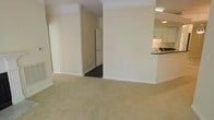 This single-story residence is designed with an open, flowing layout that connects the main living area to the surrounding spaces. The living room is finished with soft, wall-to-wall carpeting in a neutral beige tone, creating a warm and inviting atmosphere. Cream-colored walls are accented by crisp white crown molding along the ceiling, adding a classic architectural detail throughout the space.

A structural column with matching crown molding detail visually defines the transition between the living area and the adjacent kitchen. The kitchen is partially visible and features a raised breakfast bar peninsula with a dark countertop, offering a casual dining or entertaining option. Cabinetry in a light finish lines the kitchen walls, and recessed lighting illuminates the workspace.

A doorway visible from the main living area leads to an additional room or hallway, featuring a paneled white door and hardwood or dark-toned flooring beyond the threshold — a contrast to the carpeted main living space. The overall ambiance of the residence is clean, neutral, and move-in ready, with consistent finishes that provide a cohesive and functional living environment throughout.