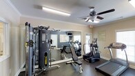 This fitness center is thoughtfully designed to accommodate a full range of workout activities within a single-level space. The room features a multi-station cable weight machine for strength training, paired with a flat workout bench, providing users with versatile resistance exercise options. On the cardio side, the space includes a treadmill, an elliptical stair climber, and additional cardio equipment visible in the mirrored wall reflection. The large floor-to-ceiling mirror along one wall enhances the sense of space while allowing users to monitor their form during exercise. A wall-mounted television is positioned above the mirror, offering entertainment during workouts. The flooring consists of durable, dark rubberized material suited for high-activity use. Crown molding and neutral beige walls give the room a polished, well-maintained appearance. A ceiling fan provides air circulation, complementing the overhead fluorescent lighting. Natural light is introduced through a window with a classic door nearby, adding brightness to the room. Fitness center guidelines are posted on the wall, ensuring users are informed of proper facility use.
