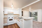 This single-floor residence offers a functional and well-organized layout with a cohesive design throughout. The kitchen is equipped with white cabinetry trimmed in warm wood accents, light-toned countertops, a coil-element electric range, a double-basin sink, and a built-in dishwasher. A peninsula counter separates the kitchen from the adjacent living and dining area, creating an open, connected flow between spaces. The hardwood flooring in the kitchen transitions to neutral wall-to-wall carpeting in the living areas.

The spacious living room is a standout feature, highlighted by a fireplace flanked by large horizontal windows that allow abundant natural light to fill the room. A sliding glass door provides direct access to an outdoor patio area with views of surrounding greenery. An additional carpeted room, suitable as a bedroom or den, includes a window with outdoor views and a baseboard heater.

The primary bedroom is generously sized, carpeted, and features natural light from a window, a baseboard heater, and an adjoining bathroom with a wide vanity, large mirror, and built-in closet with shelving. A second bathroom features warm hardwood flooring, a single-basin vanity, and clean, neutral finishes throughout.