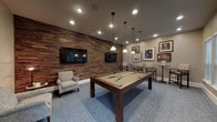 This single-floor game room is designed as a dedicated social and recreational space centered around a full-size pool table with a sage green felt surface and dark wood frame. A trio of pendant lights with white shades hangs directly above the table, providing focused task lighting complemented by recessed ceiling fixtures throughout the room. The most striking architectural feature is a full-length reclaimed wood accent wall finished in a rich, multi-toned horizontal plank pattern, lending warmth and texture to the space. A large flat-screen TV is mounted on this wall, with an additional framed display mounted nearby. Opposite the accent wall, a gallery of framed artwork is arranged on neutral beige walls, adding a curated, lounge-like aesthetic. A seating area with upholstered armchairs and a side table provides a comfortable spot for spectators. Along the windowed wall, a row of high-top bistro tables paired with tall upholstered chairs creates an additional social zone. Multiple windows fitted with horizontal blinds allow natural light to filter in, and a glass-panel door provides direct access to an outdoor area. Blue patterned carpeting covers the floor throughout, tying the room's design elements together cohesively.