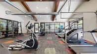 Located at 18 N Orange St in Salt Lake City, Utah, this well-equipped fitness center offers a comprehensive workout environment spread across a single level. The main exercise area is defined by soaring ceilings accented with rich, exposed wooden beams and industrial-style ductwork, lending the space a modern yet character-filled aesthetic. Large glass partition walls and oversized garage-style windows flood the interior with natural light while offering views of the surrounding greenery outdoors.

The cardio zone is generously stocked with treadmills, elliptical trainers, stair climbers, and similar machines, all arranged to maximize open floor space. A dedicated strength training area features a cable machine, a Smith machine with bench, and a tiered dumbbell rack, providing a full range of resistance training options. The flooring throughout the main gym area consists of colorful modular carpet tiles in muted tones of gray, orange, and red, adding visual interest while providing comfort underfoot.

A separate studio room with warm hardwood flooring offers additional space for stretching, yoga, or group exercise. Convenient amenities include a dual-level water fountain and restroom access. The overall ambiance is clean, bright, and functional, making this fitness center a well-rounded facility for a variety of workout routines.