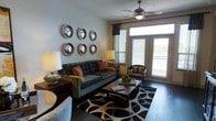 This thoughtfully designed single-floor space makes a strong first impression with its sophisticated living room. Rich hardwood-style flooring anchors the room, complemented by a bold black-and-white geometric area rug that adds visual depth and contrast. A plush, tufted gray sofa with warm burnt-orange accent pillows serves as the centerpiece, paired with a classic leather armchair and a dark wood coffee table for a balanced, inviting arrangement. A striking gallery wall of six convex round mirrors with ornate gold frames adds an artistic focal point and reflects light beautifully throughout the room. A ceiling fan with a built-in light fixture overhead provides both comfort and ambient illumination. Large windows dressed with slatted blinds and dark curtain panels flood the space with natural light, while a set of glass-paned French doors opens to a private outdoor balcony, offering pleasant views of the surrounding greenery. Table lamps on either side of the seating area provide warm, layered lighting. The overall ambiance is warm yet contemporary, blending classic decorative touches with modern furnishings to create a welcoming and functional living environment.