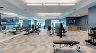 Located at 265 Communication Way in Hyannis, Massachusetts, this single-floor fitness facility offers a comprehensive wellness environment designed for both individual training and group exercise. The main gym area is expansive and filled with an extensive array of strength and cardio equipment, including cable machines, weight benches, resistance machines, ellipticals, and more. Bold teal accent walls, large windows that invite natural light, and a distinctive chevron-patterned floor tile give the space a modern, energizing aesthetic. Overhead, flush panel ceilings with recessed fluorescent lighting ensure the room is brightly and evenly illuminated throughout. A wall-mounted flat-screen television and a small refreshment station add convenience for members during their workouts. A dumbbell rack near the entrance provides quick access to free weights. Adjacent to the main floor is a dedicated multipurpose studio, separated by a large mirrored wall that enhances the sense of space. This room features light wood-look flooring, a wall-mounted flat-screen display, rows of padded chairs for group sessions, and storage for step platforms and exercise mats. A wall-mounted yoga mat holder and colorful artwork contribute to the welcoming, functional character of the studio. Together, the areas form a well-rounded fitness destination suitable for a wide range of exercise programs.