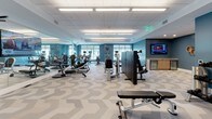 Located at 265 Communication Way in Hyannis, Massachusetts, this single-floor fitness facility offers a comprehensive wellness environment designed for both individual training and group exercise. The main gym area is expansive and filled with an extensive array of strength and cardio equipment, including cable machines, weight benches, resistance machines, ellipticals, and more. Bold teal accent walls, large windows that invite natural light, and a distinctive chevron-patterned floor tile give the space a modern, energizing aesthetic. Overhead, flush panel ceilings with recessed fluorescent lighting ensure the room is brightly and evenly illuminated throughout. A wall-mounted flat-screen television and a small refreshment station add convenience for members during their workouts. A dumbbell rack near the entrance provides quick access to free weights. Adjacent to the main floor is a dedicated multipurpose studio, separated by a large mirrored wall that enhances the sense of space. This room features light wood-look flooring, a wall-mounted flat-screen display, rows of padded chairs for group sessions, and storage for step platforms and exercise mats. A wall-mounted yoga mat holder and colorful artwork contribute to the welcoming, functional character of the studio. Together, the areas form a well-rounded fitness destination suitable for a wide range of exercise programs.